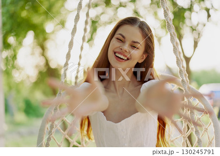 Young woman smiling warmly while sitting in a swing outdoors, wearing a white dress, surrounded by greenery, evoking joy and happiness in a serene summer setting 130245791