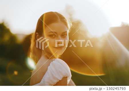 Young woman in a sunlit field, wearing a white dress, smiling softly, conveying joy and serenity against a warm golden background 130245793