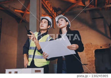 Construction manager and engineer dressed in orange work vests and hard helmets explore construction documentation on the building site near the steel frames 130245905
