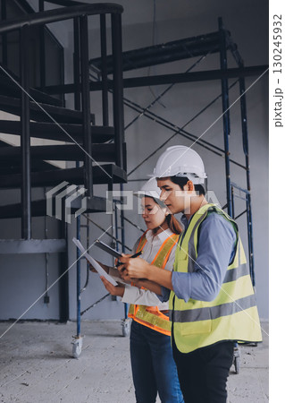 Construction manager and engineer dressed in orange work vests and hard helmets explore construction documentation on the building site near the steel frames 130245932