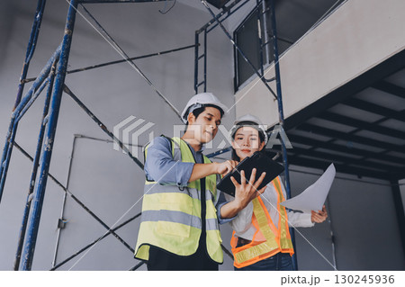 Construction manager and engineer dressed in orange work vests and hard helmets explore construction documentation on the building site near the steel frames Construction manager and engineer dressed in orange work vests and hard helmets explore construction documentation on the building site near the steel frames 130245936