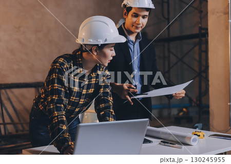 Construction manager and engineer dressed in orange work vests and hard helmets explore construction documentation on the building site near the steel frames Construction manager and engineer dressed in orange work vests and hard helmets explore construction documentation on the building site near the steel frames 130245955
