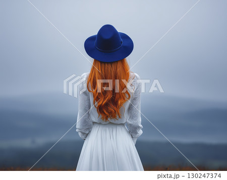Elegant woman stands serenely, back to camera, gazing towards hazy landscape, her vibrant hair striking contrast against cool tones of misty mountains 130247374