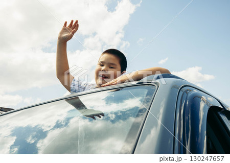 happy boy smiling and waving hand while looking out from car sunroof, blue sky with white clouds, joyful childhood moment of summer travel and family road trip, family, vacation, lifestyle 130247657