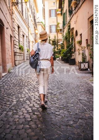 Woman with hat and backpack walking along a narrow cobblestone street in Rome surrounded by historic buildings and greenery Woman with hat and backpack walking along a narrow cobblestone street in Rome surrounded by historic buildings and greenery 130247978