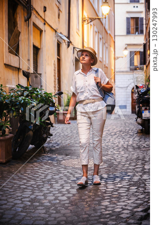 Middle-aged woman in light clothes and straw hat walking along a narrow cobblestone street in Rome, looking up at historic architecture illuminated by evening lamps Middle-aged woman in light clothes and straw hat walking along a narrow cobblestone street in Rome, looking up at historic architecture illuminated by evening lamps 130247993