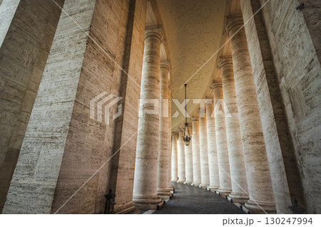 Curved colonnade with massive stone pillars and hanging lanterns in St. Peter's Square, Vatican City, creating a grand architectural perspective 130247994