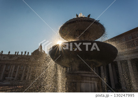 Fountain in St. Peter's Square with sunlight shining through cascading water, creating a dramatic silhouette of Vatican architecture 130247995