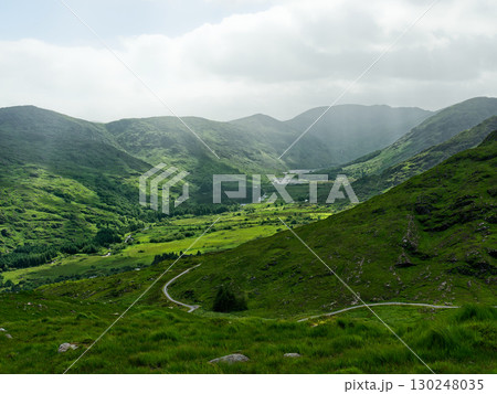 A landscape view shows green hills under an overcast sky. A winding road cuts through the lush valley floor in Gap of Dunloe, County Kerry, Ireland. A landscape view shows green hills under an overcast sky. A winding road cuts through the lush valley floor in Gap of Dunloe, County Kerry, Ireland. 130248035