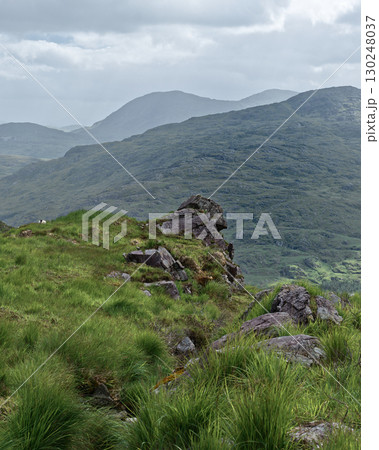 A serene landscape unfolds, revealing the lush green hills and rugged rock formations within the Gap of Dunloe. Cloudy skies provide a soft light over the mountainous terrain in Ireland. A serene landscape unfolds, revealing the lush green hills and rugged rock formations within the Gap of Dunloe. Cloudy skies provide a soft light over the mountainous terrain in Ireland. 130248037