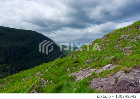 The rolling, grassy hills meet in a valley. Scattered rocks dot the grassy landscape under a cloudy sky. This is the Gap of Dunloe in County Kerry, Ireland during summertime. 130248042