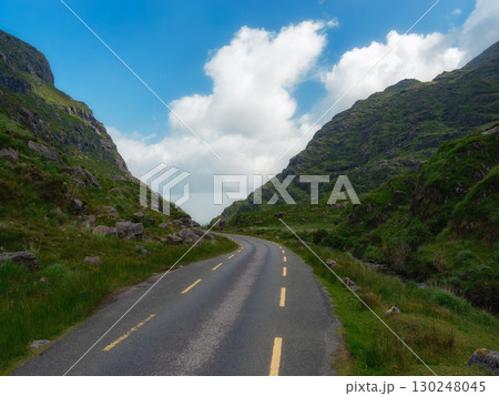 A winding road cuts through a lush green valley, nestled between imposing mountains under a vibrant blue sky dotted with fluffy white clouds. The scene evokes a sense of adventure and tranquility. 130248045