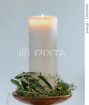 Large white pillar candle burns softly atop a wooden holder adorned with fresh greenery. The simple yet elegant arrangement brings a sense of calm and tranquility to the setting. 130250491