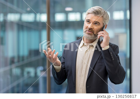 A confident man in a suit having a phone call, standing in a contemporary office setting. A confident man in a suit having a phone call, standing in a contemporary office setting. 130250817