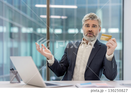 Mature businessman appears concerned while holding a bank card and working on his laptop in an office. He seems troubled, conveying potential issues with finance, payment, or online security. 130250827