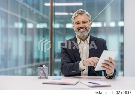 Confident middle-aged professional using a tablet at a modern office desk with joy. Portrays thoughtfulness, technology use, and the workplace environment, conveying professionalism Confident middle-aged professional using a tablet at a modern office desk with joy. Portrays thoughtfulness, technology use, and the workplace environment, conveying professionalism 130250832