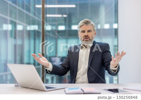 A mature businessman gesturing with frustration while seated at a desk in a contemporary office environment. The image conveys emotions of confusion and miscommunication in a professional workspace. 130250848
