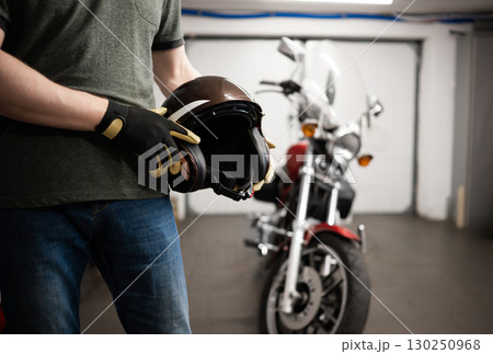 Man Holds A Protective Moto Helmet Against A Classic Motorcycle In A Garage Man Holds A Protective Moto Helmet Against A Classic Motorcycle In A Garage 130250968