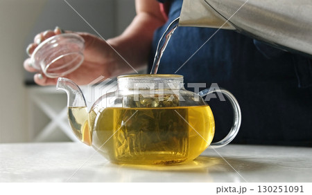 Girl Pouring Hot Water Into A Clear Teapot Filled With Fresh Green Tea On A Kitchen Table Girl Pouring Hot Water Into A Clear Teapot Filled With Fresh Green Tea On A Kitchen Table 130251091