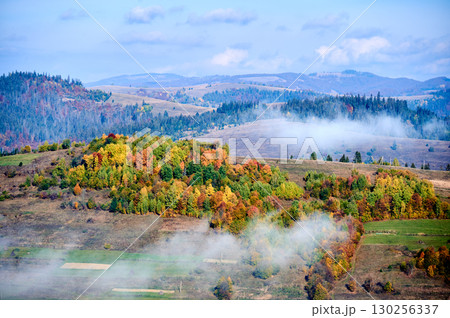 Picturesque view of hillside adorned with vibrant autumn foliage, ranging from rich greens to fiery reds and golden yellows. Wisps of fog drift across landscape under bright blue sky. 130256337