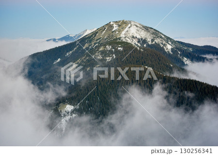 Breathtaking photograph showcases majestic mountain peak, partially covered with snow and surrounded by dense pine forests. Mist and clouds weave through valleys under clear blue sky. Breathtaking photograph showcases majestic mountain peak, partially covered with snow and surrounded by dense pine forests. Mist and clouds weave through valleys under clear blue sky. 130256341