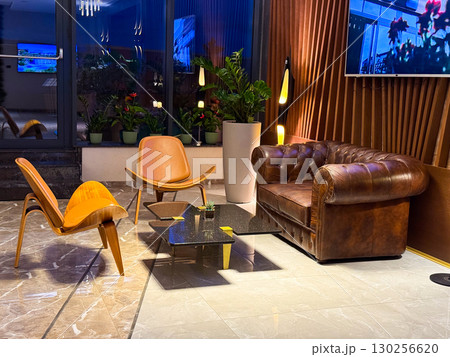 Elegant hotel lounge with leather sofa, wooden chairs and marble flooring. Retro inspired seating arrangement emphasizing hospitality, comfort and welcoming atmosphere. 130256620