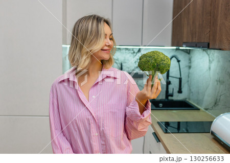 Smiling young woman in pink shirt holding and smelling fresh broccoli in kitchen, enjoying healthy lifestyle and organic food. 130256633