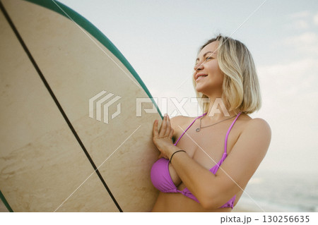 Young blonde woman in a purple bikini holding a surfboard, smiling and enjoying the beach. Ocean waves in the background, summer lifestyle and carefree vacation vibes. Young blonde woman in a purple bikini holding a surfboard, smiling and enjoying the beach. Ocean waves in the background, summer lifestyle and carefree vacation vibes. 130256635