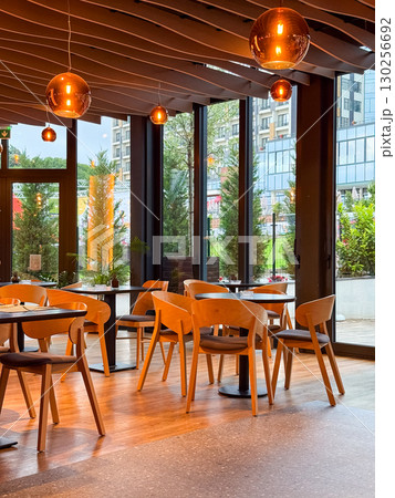 Bright restaurant hall with tables and chairs arranged near the glass doors leading outside. Hospitality, service, lifestyle and dining atmosphere in modern interior. Bright restaurant hall with tables and chairs arranged near the glass doors leading outside. Hospitality, service, lifestyle and dining atmosphere in modern interior. 130256692
