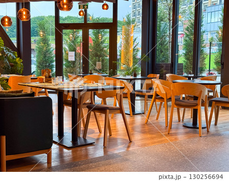 Bright restaurant hall with tables and chairs arranged near the glass doors leading outside. Hospitality, service, lifestyle and dining atmosphere in modern interior. 130256694