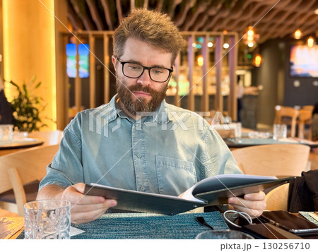 Man sitting at a restaurant table reading menu and using smartphone in cozy modern interior. Lifestyle, leisure, and dining experience symbolizing hospitality, communication, and contemporary food 130256710