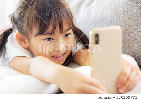 ソファに寝転んでスマホを見る女の子 130259772