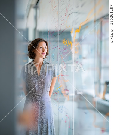businesswoman standing near glass wall covered in graphs and schedules 130261197