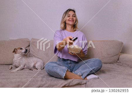 Happy young woman watching TV and smiling, holding popcorn bowl, sitting on sofa with cute dog. Cozy home evening, relaxing time with pet and favorite show. 130262955