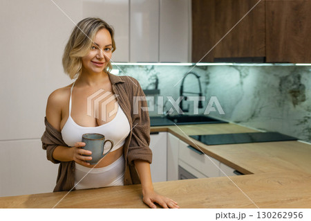 Modern kitchen: Young woman in a white crop top and open shirt stands with a mug in hand, enjoying a peaceful morning at home, relaxed and smiling. Modern kitchen: Young woman in a white crop top and open shirt stands with a mug in hand, enjoying a peaceful morning at home, relaxed and smiling. 130262956