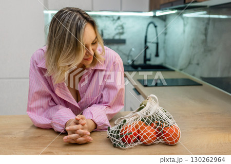 Smiling woman in pink shirt leaning on kitchen counter next to mesh bag with fresh vegetables. Healthy lifestyle and eco-friendly food shopping at home. 130262964