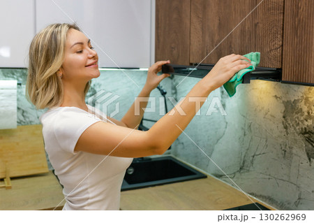 Smiling woman cleaning kitchen furniture with cloth, enjoying housework. Young adult wiping wooden cabinet in modern bright kitchen interior, daily cleaning routine at home. 130262969