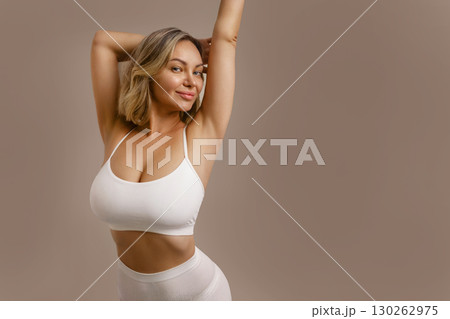 Young beautiful woman in white lingerie, smiling and touching her body, showing natural beauty, breast care, body positivity and healthy lifestyle on beige background. 130262975