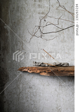 Burning incense stick on a rustic holder with ash, placed against a textured concrete wall with dry branches and old wood. Minimalist still life in wabi-sabi style. 130263194
