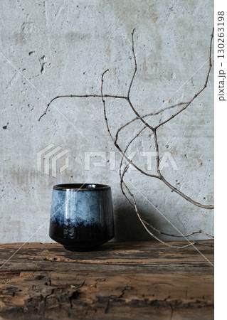 Minimalist still life with a dark ceramic vase and dry branch on rustic wood against a textured concrete wall. Wabi-sabi style composition Minimalist still life with a dark ceramic vase and dry branch on rustic wood against a textured concrete wall. Wabi-sabi style composition 130263198