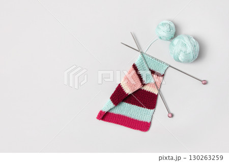 Brown beige vinous and turquoise piece of striped knitting with knitting needles and balls of thread on gray background. Homemade needlework and knitting of warm, clothes and toys. Concept creativity 130263259