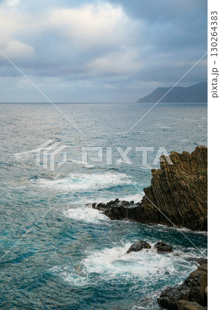 A view of sea waves crashing against a rocky cliff, with another island A view of sea waves crashing against a rocky cliff, with another island 130264383