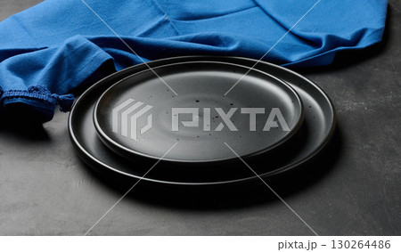 Empty black ceramic plates stacked on dark background with blue linen napkin. Minimalist food still life. 130264486