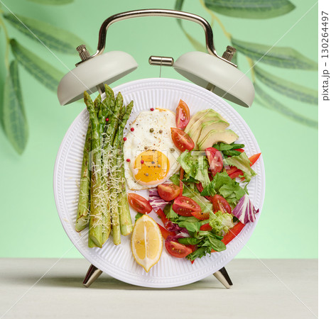 Plate with asparagus, fried eggs and vegetable salad, stylized as an alarm clock on a green background. 130264497