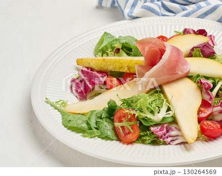 Close up of a gourmet salad with prosciutto, pear slices, cherry tomatoes and fresh mixed greens served on a white ribbed plate. Close up of a gourmet salad with prosciutto, pear slices, cherry tomatoes and fresh mixed greens served on a white ribbed plate. 130264569