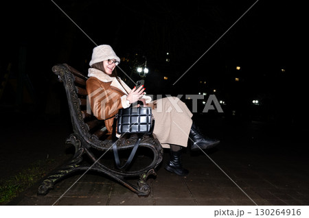 Woman enjoying a quiet evening with her phone on a park bench under the moonlight, surrounded by distant city lights and the tranquility of the night 130264916