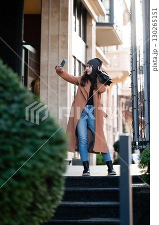 Charming city stroll captured in vibrant sunlight as a young woman enjoys a moment with her phone in a stylish coat and hat during an afternoon outing 130265151