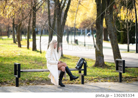 Bright autumn day in the park featuring a thoughtful young woman sitting on a bench while looking at her phone and enjoying the beautiful surroundings Bright autumn day in the park featuring a thoughtful young woman sitting on a bench while looking at her phone and enjoying the beautiful surroundings 130265334