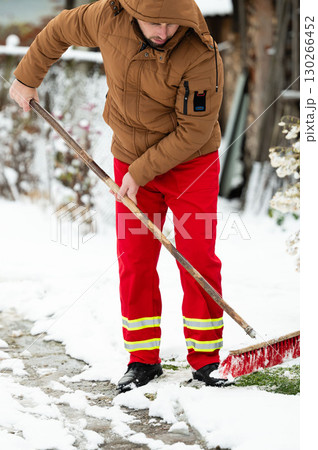 Snowy day in the city as a dedicated worker clears the path with a shovel, showcasing determination in bright red attire against the winter backdrop 130266452