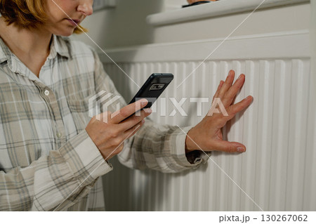 Woman holding smartphone next to heating thermostat, increasing heating costs Woman holding smartphone next to heating thermostat, increasing heating costs 130267062